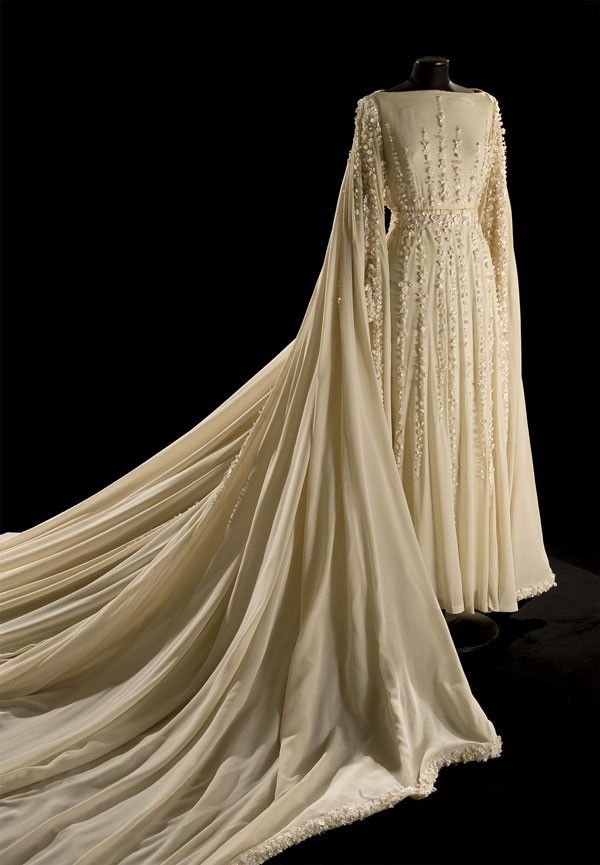 a creamcolored wedding dress with long sleeves and a long train is displayed on a mannequin against a black background. the dress features delicate embellishments throughout. the fabric is flowing and elegant.