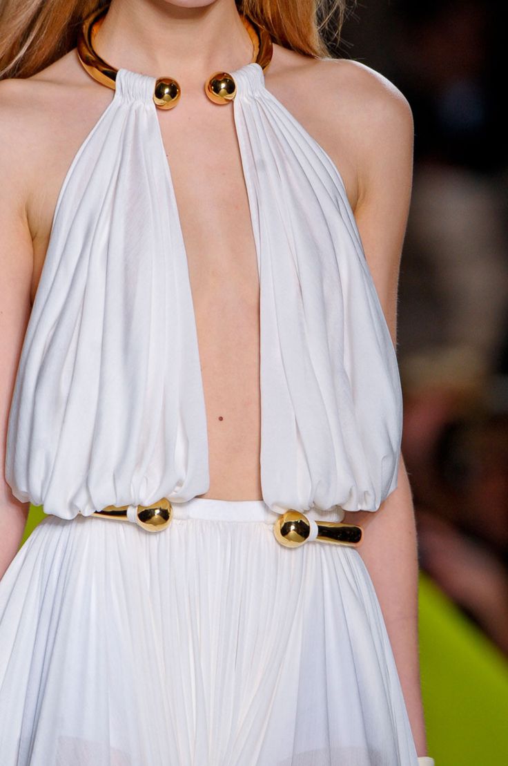 closeup view of a model on a runway wearing a stunning white halter neck dress. the dress is pleated and features a minimalist design with gold accents. a wide gold necklace with spherical ends sits at the neckline, and a matching gold belt cinches the waist. the model's skin is fair, and her hair is light brown, pulled back. the background is blurred, suggesting a runway setting. the overall impression is one of elegance and high fashion.