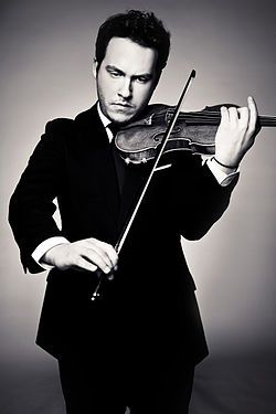 a black and white portrait of a man in a suit playing the violin. he is standing and holding the violin in the traditional manner, with his left hand on the fingerboard and his right hand on the bow. he is looking down at the violin with a serious expression on his face. the background is a plain gray.
