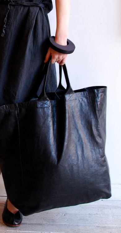a woman in a black dress holding a large black leather tote bag with a thick black bracelet on her wrist and wearing black shoes.