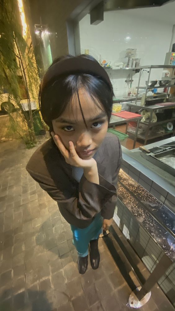 young girl is taking a selfie in a restaurant. she is wearing a brown blazer, blue jeans, black shoes, and a black headband. the image is taken from a high angle, giving a bird's eye view. a fisheye lens is used, creating a distorted perspective. the restaurant is dimly lit with artificial lighting.