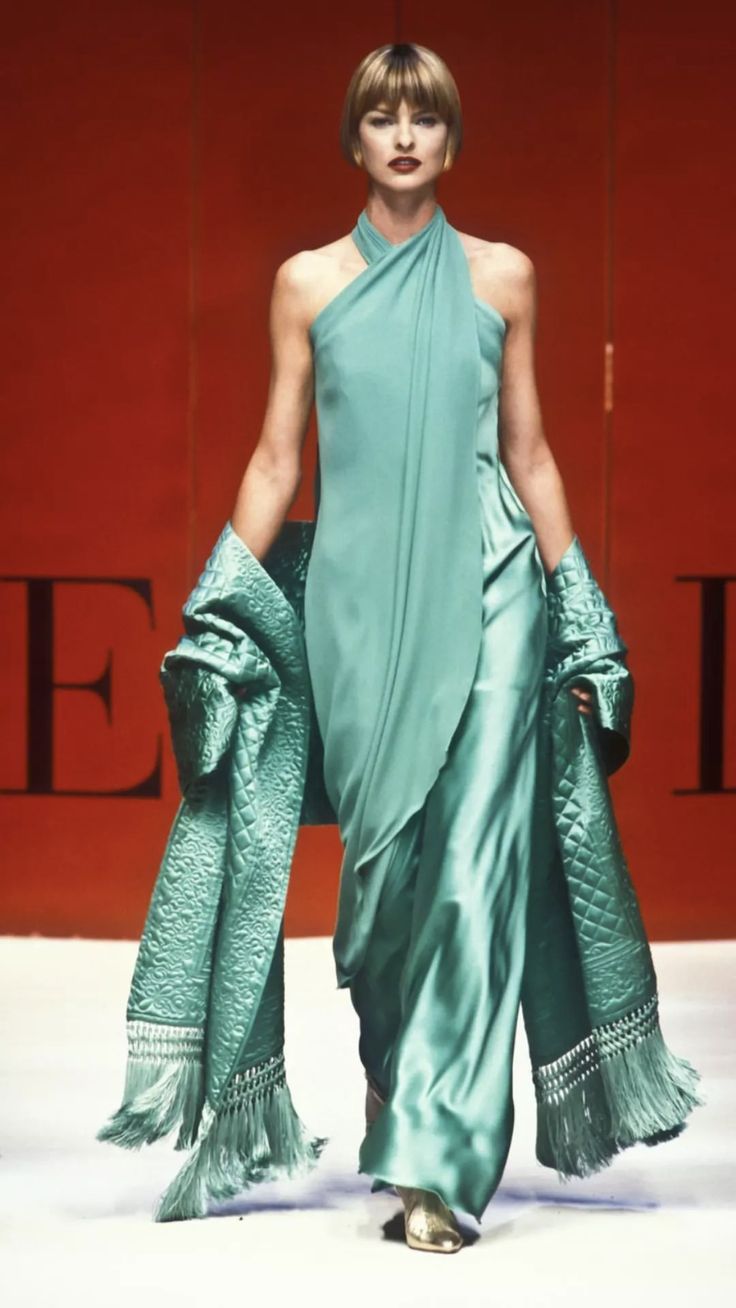 a fashion model walks down a runway in a long, teal green dress with a matching fringed shawl. she has short blonde hair and is wearing high heeled shoes. the background is a red wall.