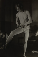 a black and white photograph showing a male ballet dancer backstage, putting on his tights. the dancer is shirtless and is in the process of pulling up his white tights. he is standing with one leg extended in a ballet pose. the background is dark and blurred.