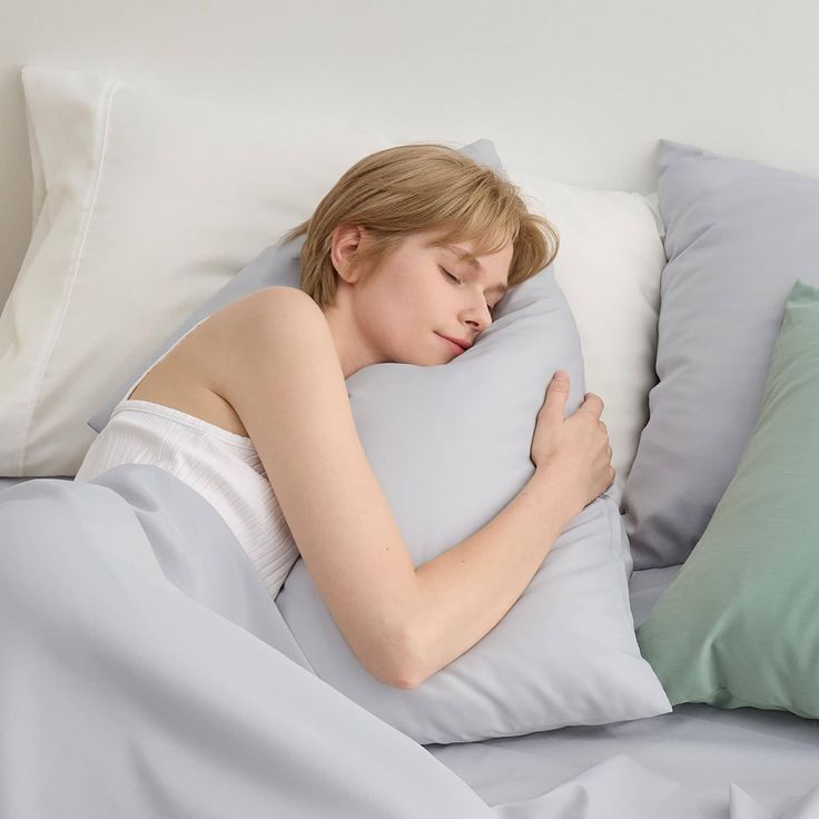 a woman with short blonde hair sleeps peacefully in a bed with white and light green pillows and light blue sheets. she's hugging a light blue pillow. the image evokes a sense of calm and relaxation.