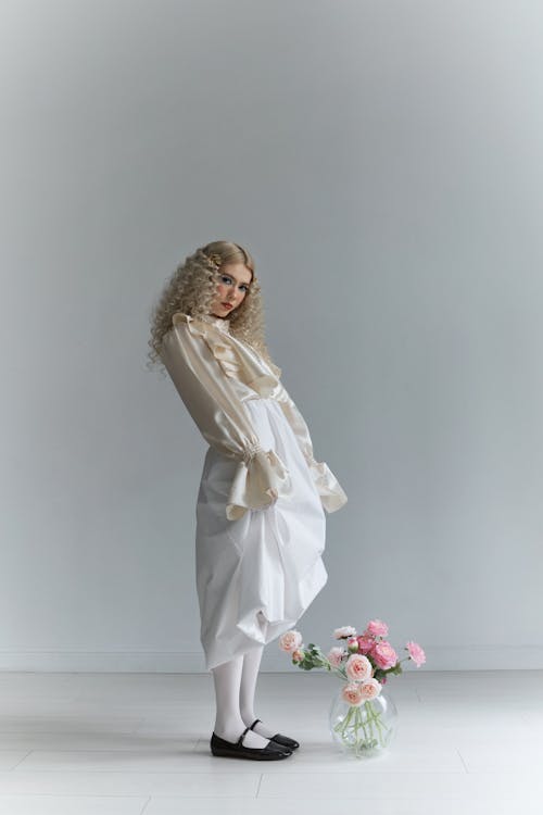a woman with blonde, curly hair is standing indoors, posing in front of a grey backdrop. she is wearing a long, lightcolored dress with ruffled sleeves, white tights, and black ballet shoes. next to her is a vase filled with pink and peachcolored flowers. she is looking directly at the camera.