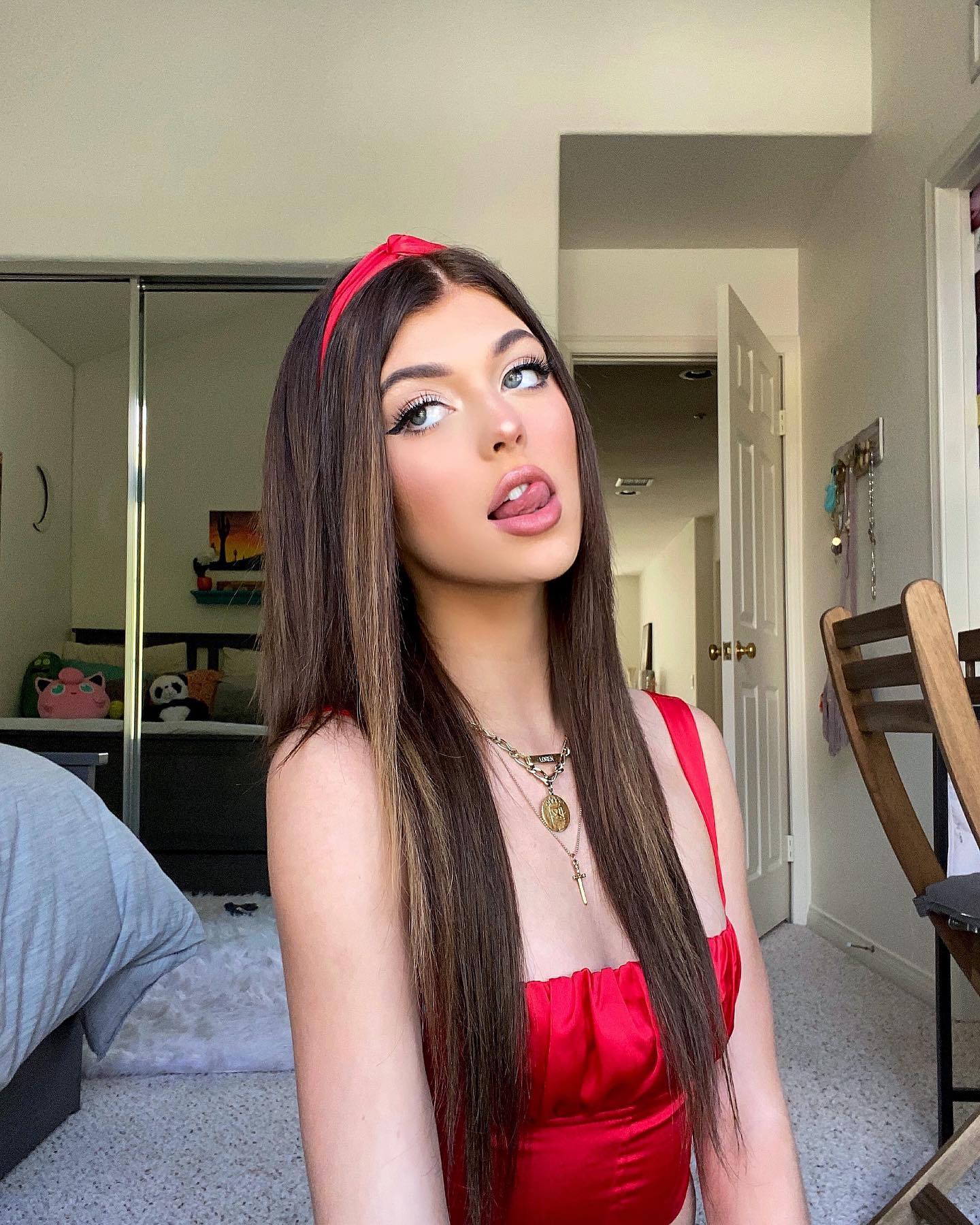 a woman with long brown hair, a red headband, and a red top is sticking her tongue out while looking to her left. she is wearing a necklace and is in a bedroom. there is a mirror, a bed, a pillow, a doorway, and a chair in the background.