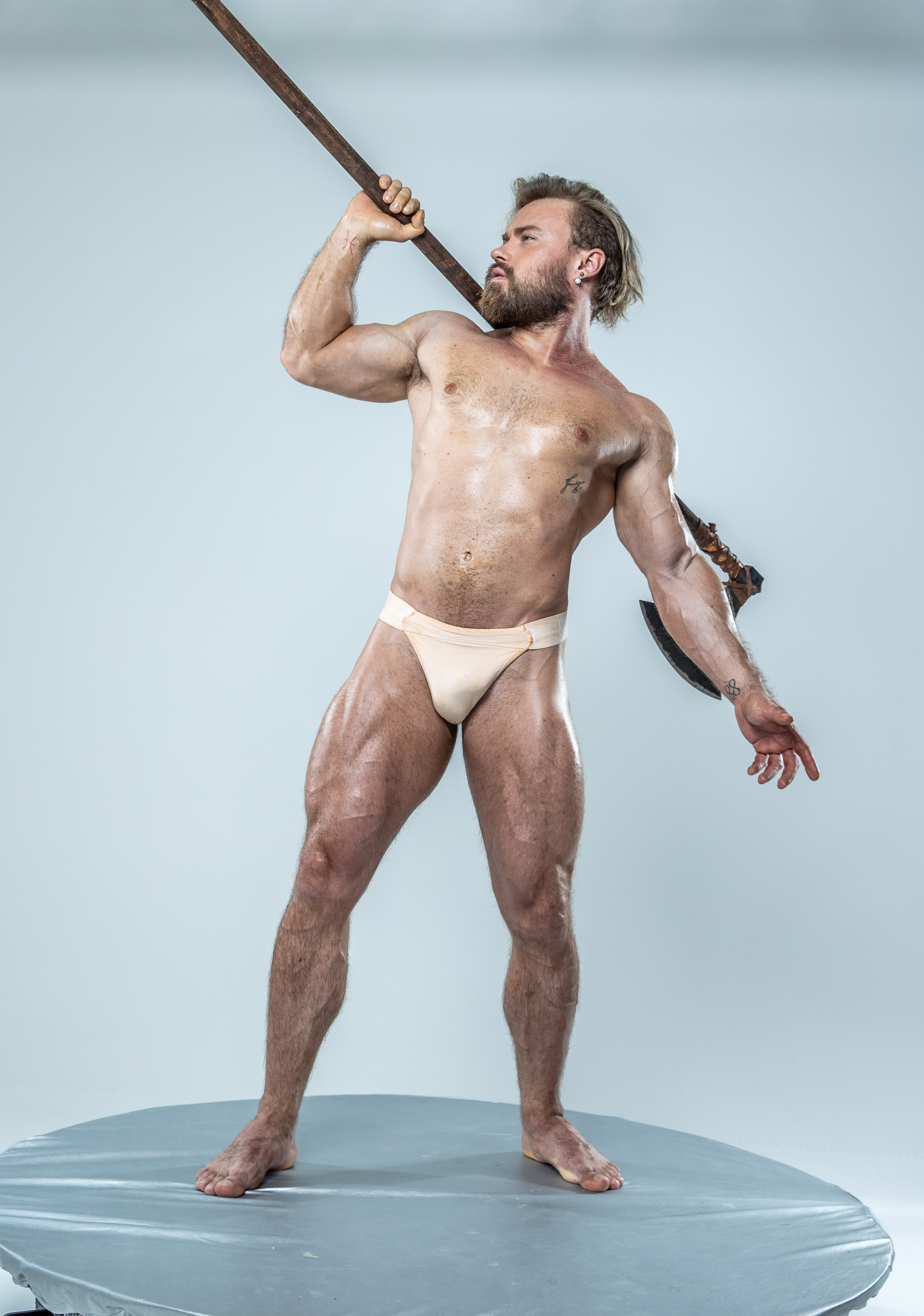 a muscular, bearded man with a toned physique stands on a round platform in a bright studio. he is wearing only underwear and is holding a long axe over his shoulder, his body glistening with sweat. the pose emphasizes his strength and power, evoking imagery of a warrior or mythological figure.