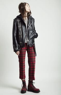 a woman wearing a black leather jacket with pins, red and black plaid pants, and black boots poses for a photo. she is looking to the right of the frame.