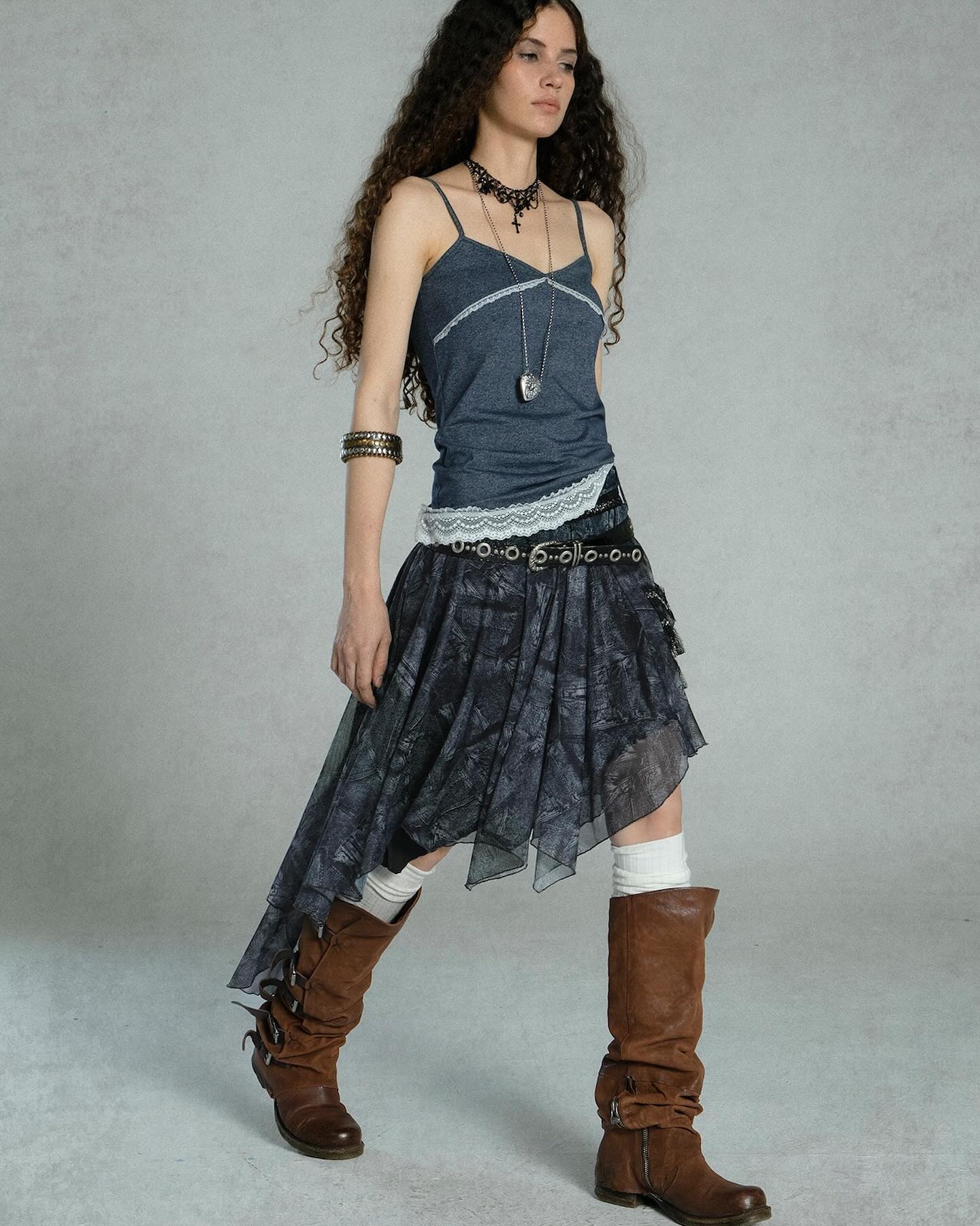 fulllength studio shot of a woman, possibly a model, dressed in a bohemianstyle outfit. she is wearing a dark gray sleeveless top with lace trim, a dark gray and black asymmetrical skirt, brown leather boots, and several accessories including a necklace and bracelets.  her long, curly brown hair is loose around her shoulders.  the background is a simple, light gray. the overall style is casual and slightly grunge with a bohemian flair.