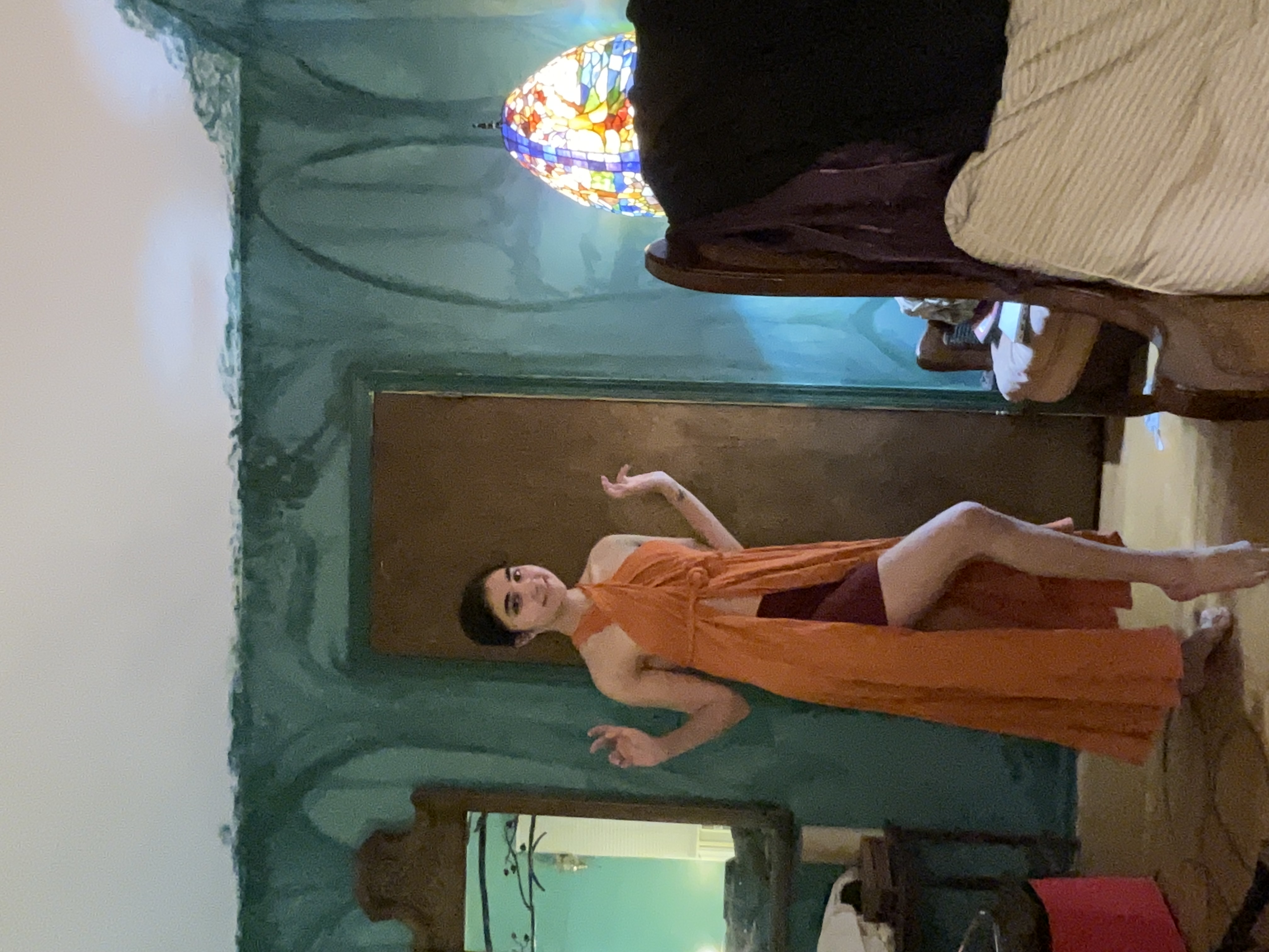 a young woman with shoulderlength dark hair is standing in a bedroom. she's wearing a long, orange wrap dress with a high slit, showing off a burgundy colored underskirt. the dress has a halter neckline. she's smiling and striking a pose with her arms slightly outstretched. the room's walls are painted a teal or dark green color with uneven brushstrokes, suggesting it's unfinished or designed to look rustic. there is a vintage stainedglass lamp, and an antiquestyle mirror with a wooden frame. a partially visible upholstered chair and bed are also in the background. the overall aesthetic suggests a bohemian or eclectic style.