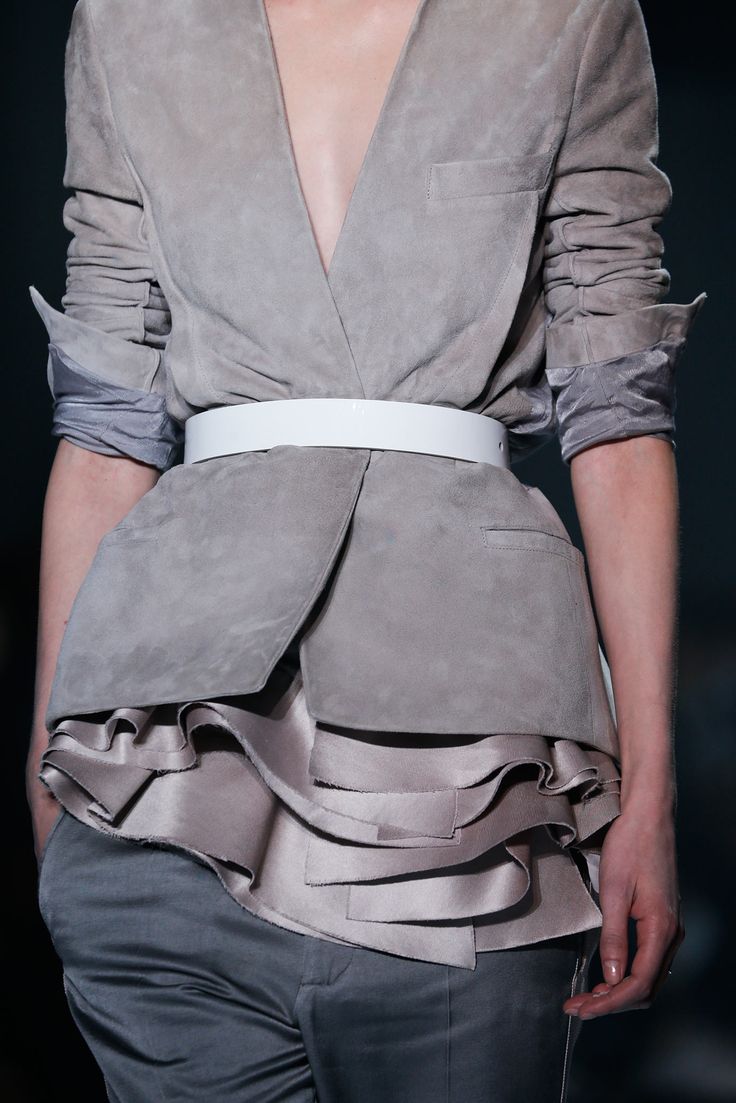 a female model in a gray suede blazer with a white belt and ruffles at the bottom. the jacket is unbuttoned and the woman is standing with one hand on her hip, the other hand is resting on the side of her body, and her head is turned away from the camera.