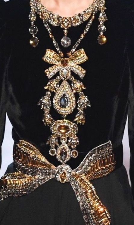 closeup view of a woman's black velvet dress adorned with an elaborate, multilayered necklace and matching jeweled bow at the waist. the necklace features a large, central pendant with gold, silver, and crystal embellishments. the bow at the waist is similarly decorated with gold and silver beads and crystals. the overall aesthetic is opulent and extravagant, suggestive of highfashion or runway attire.  the dress and jewelry display intricate details and exceptional craftsmanship.