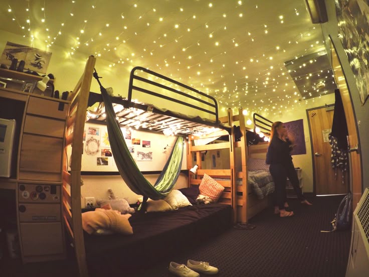 this image shows a dorm room with bunk beds. a hammock is strung between the beds. string lights decorate the ceiling. a woman is standing in the room. there are pillows on the beds and posters on the walls. a heater is visible in the corner.