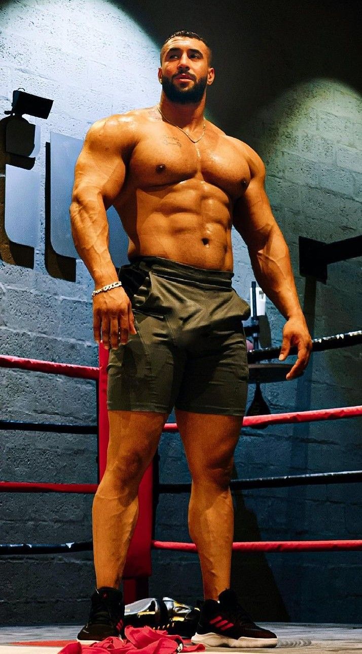 the image shows a muscular, shirtless man standing in a boxing ring.  he's exhibiting a very defined physique with a sixpack and welldeveloped muscles. he's wearing dark athletic shorts and sneakers. the background is dark and moody, with a spotlight illuminating the man. the overall impression is one of strength, fitness, and athleticism. a pair of boxing gloves are visible on the floor near his feet.