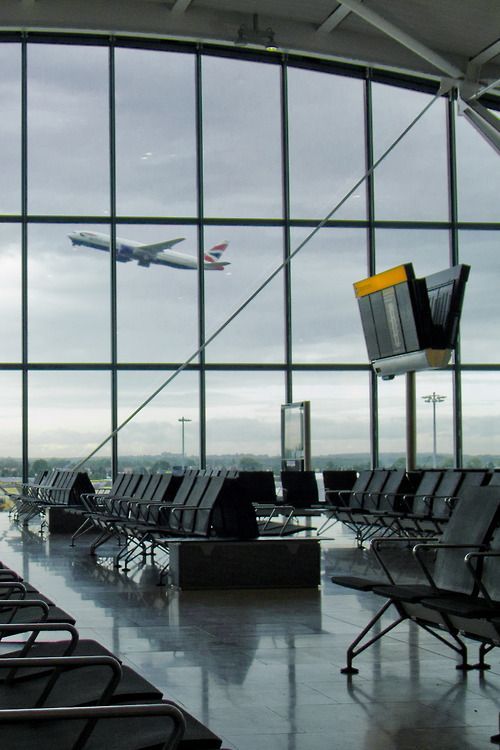 the image shows the interior of a modern airport terminal.  a large panoramic window dominates the background, offering a view of a british airways boeing 747 airplane taking off. the foreground shows rows of empty, modern airport waiting chairs arranged neatly on a highly polished floor.  a flight information display (fid) is visible on the right, and a portion of another is glimpsed behind the chairs. the overall impression is one of sleek, contemporary design and a sense of anticipation associated with air travel. the sky outside is overcast.