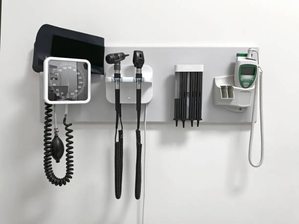 a white wall with medical equipment mounted on it. there is a blood pressure monitor, otoscope, stethoscope, and a thermometer.