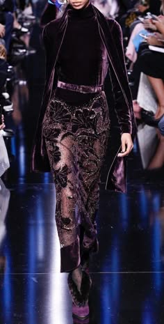 a female model wearing a long purple velvet dress over a sheer patterned skirt walks the runway. she has her hair pulled back and is wearing makeup that complements the purple dress. the background is filled with a blurred view of the audience.