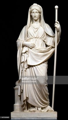 a fulllength marble statue of a woman in roman attire, standing contrapposto and holding a staff in her right hand. she wears a draped gown and a veil covers her head. the statue is positioned on a pedestal, set against a black background.