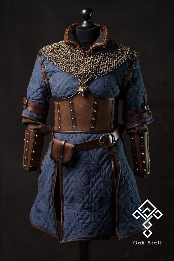 a mannequin is wearing a blue quilted tunic with brown leather trim, a brown leather corset with metal studs, chainmail around the neck, and brown leather gauntlets. the logo for oak stall is in the bottom right corner.