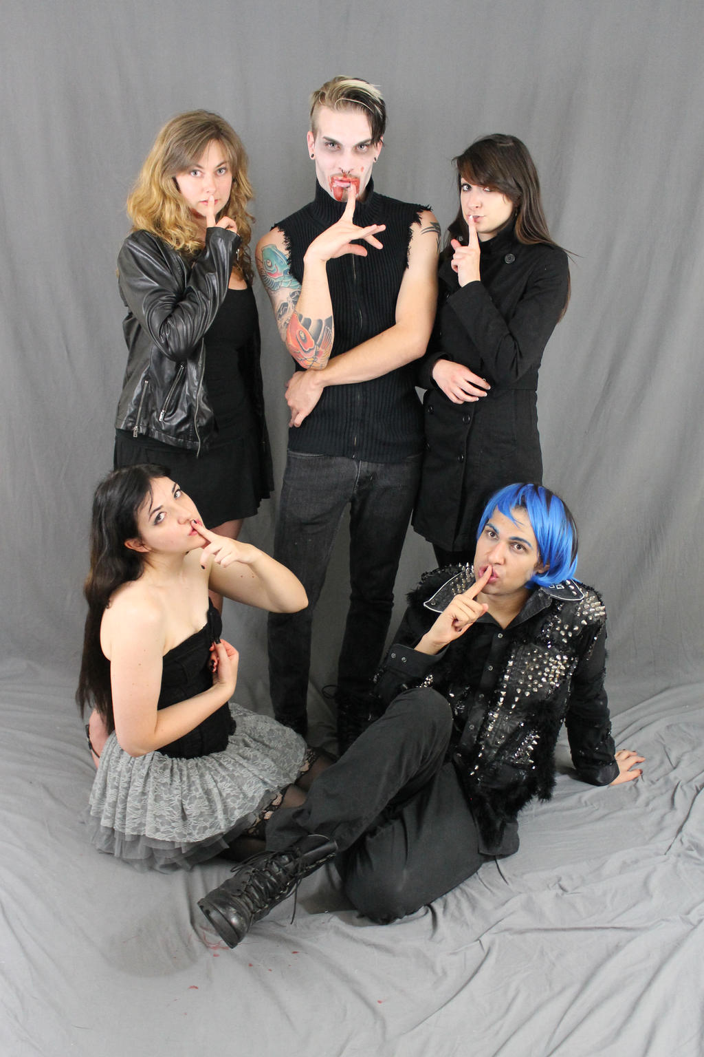 group photo of five young adults, styled in a gothic/alternative aesthetic, posing against a gray backdrop.  the central figure is a man with blonde hair and vampirethemed makeup. the others are two women in black leather jackets and skirts, a woman in a black tulle skirt, and a man with blue hair and a studded black shirt.  all are making a "shhh" gesture with their fingers to their lips.  the overall mood is dark, mysterious, and slightly theatrical.