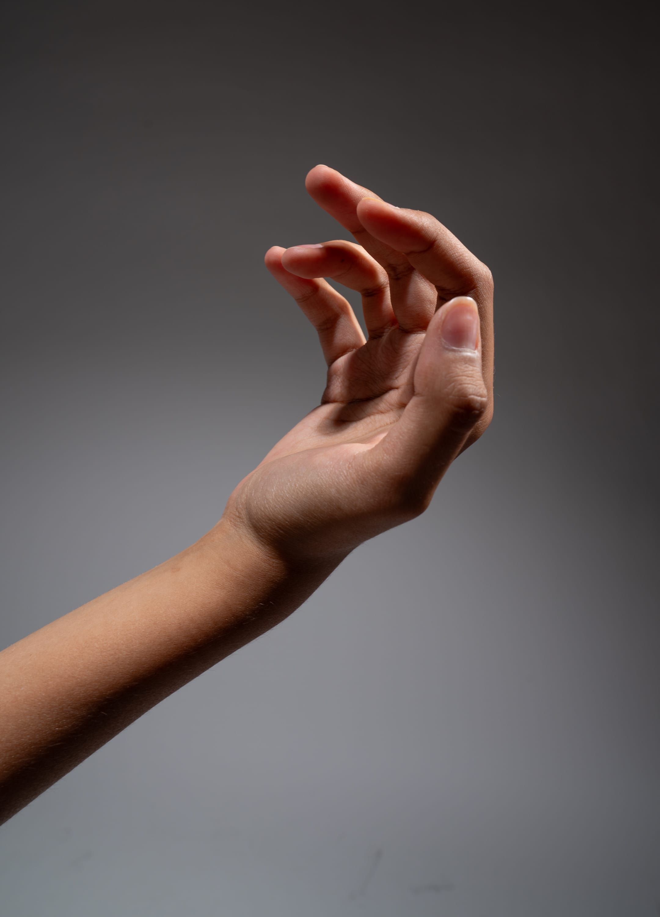 a hand is extended, its fingers slightly curled inward. the lighting is soft and directional, illuminating the hand and arm against a neutral grey background.