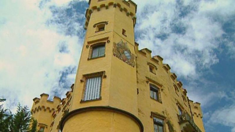 a low angle view of a yellow castle tower against a blue sky with white clouds. the tower is made of stone with multiple windows, some with white coverings, and features a sundial on its side.  the image showcases the castle's architectural details.