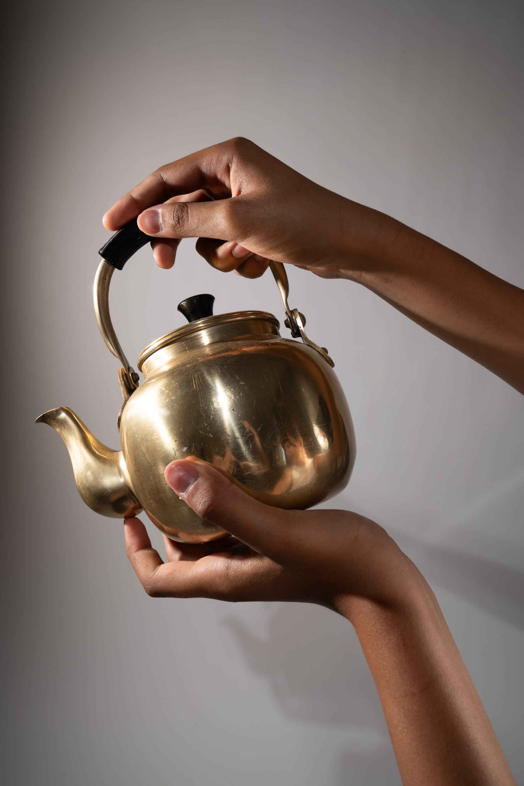 two black hands are holding a gold tea kettle. the kettle is made of a metallic material and has a black handle. the hands are positioned in a way that suggests the person is about to pour tea. the background is a plain, light gray wall. the lighting is soft and even, creating a gentle highlight on the kettle's surface. the overall composition is minimalist and elegant.