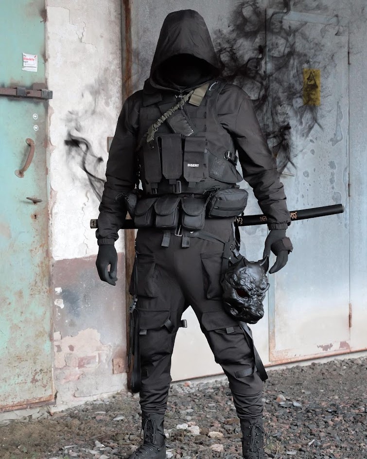 a person in a black hooded jacket and face mask stands in front of a concrete wall, their tactical gear and sword on display. they are wearing black cargo pants, boots, and gloves, and their face is obscured by the hood. the person is posing with a katana sword and a demon mask hanging on their belt. the photo is styled to look like a cyberpunk action scene, with dark shadows and a menacing atmosphere.
