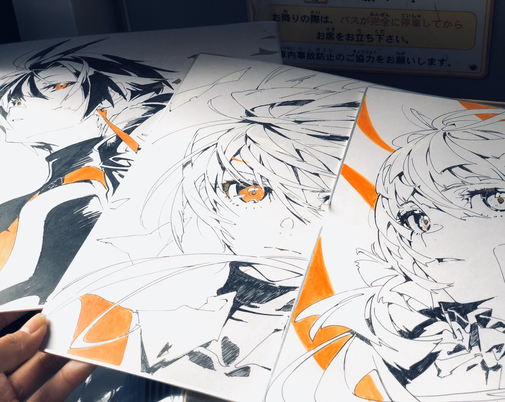 n artist holds up three impressive, nearly finished anime character sketches against the backdrop of a public transit bus interior. the black and white drawings, accented with vibrant orange highlights, hint at a dynamic and stylized art style. the bus's safety notice peeks out from behind the artwork, adding a layer of everyday context to the artistic display.