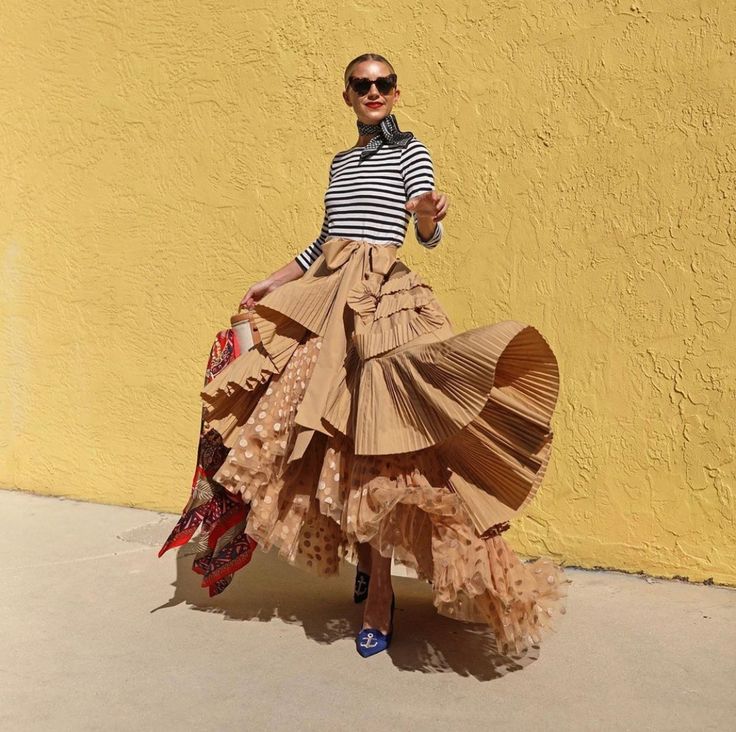 a woman stands outdoors against a bright yellow wall, wearing a stylish ensemble. she sports a black and white striped shirt, accessorized with a patterned scarf and sunglasses. her statement piece is a layered skirt in varying textures and shades of beige/tan. the skirt features pleats, ruffles, polka dots, and a tulle overlay, creating a voluminous and dynamic silhouette. she completes her look with blue shoes adorned with an anchor symbol.