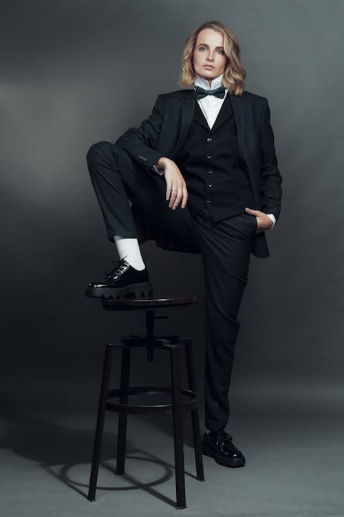 a full shot of a woman model confidently posing in a stylish black tuxedo suit. she is wearing a crisp white shirt underneath and a black bow tie. her blonde hair is styled in a short, wavy bob. one leg is casually resting on a black metal stool, and she wears black oxford shoes.  the background is a dark gray, providing a stark contrast to her attire. the overall impression is one of elegance and sophistication with an androgynous edge.
