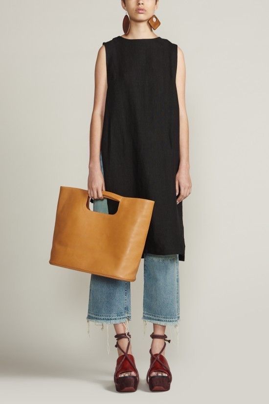 a fulllength studio shot features a woman modeling a chic, contemporary outfit. she is adorned with prominent geometric brown earrings, wearing a simple black sleeveless dress that falls to midthigh. below the dress, she sports light blue denim jeans with a slightly cropped, wideleg style, complete with a fringed hem for a touch of edginess. on her feet, she has on red square toe platform sandals that lace up the ankles, adding height and a pop of color to the ensemble. in her left hand, she carries a structured brown leather handbag with square handles, complementing the overall color palette and adding a touch of sophistication. the background is a plain light beige color, ensuring that the focus remains on the fashion and the model.