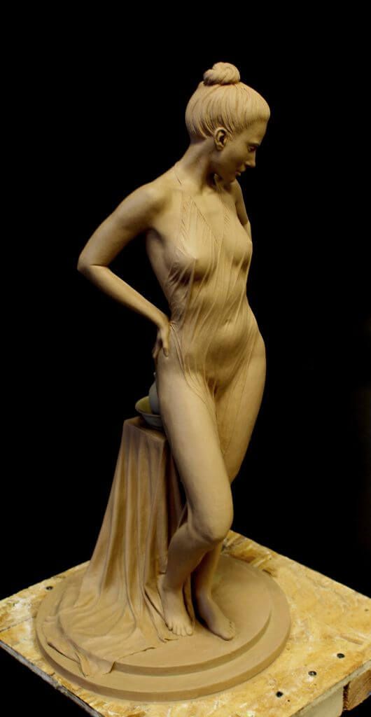 a clay sculpture of a nude woman standing with her hand on her hip. she has her hair tied up and is looking down. the sculpture is very detailed and realistic, showing the curves of the woman's body. the sculpture is standing on a wooden platform. the background is black.