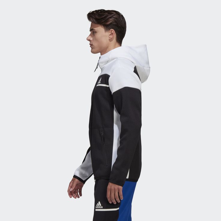 a man is wearing a black and white adidas jacket. the jacket has a white zipper, and the model is wearing blue pants. he has short brown hair and is looking to his left side. the background is white.
