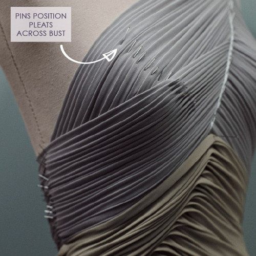 a closeup of a grey and beige pleated dress being designed on a mannequin. the grey pleats are pinned in place across the bust area, and there is a callout box with an arrow pointing to the pins, stating "pins position pleats across bust." the lower part of the dress is in a tan or beige color and also features pleats.