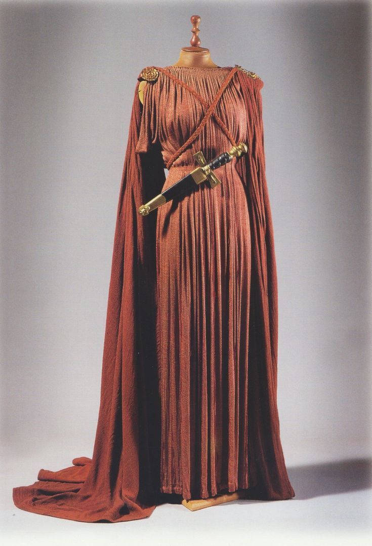 full shot of a terracottacolored ancient greekstyle dress displayed on a mannequin. the dress is a long, pleated garment with a shortsleeved bodice and a long, flowing overdress or himation in a similar color. a dagger is sheathed at the waist. the fabric is thick and appears to be made of a heavy material. the background is a neutral light gray. the image is welllit and shows the details of the costume clearly.