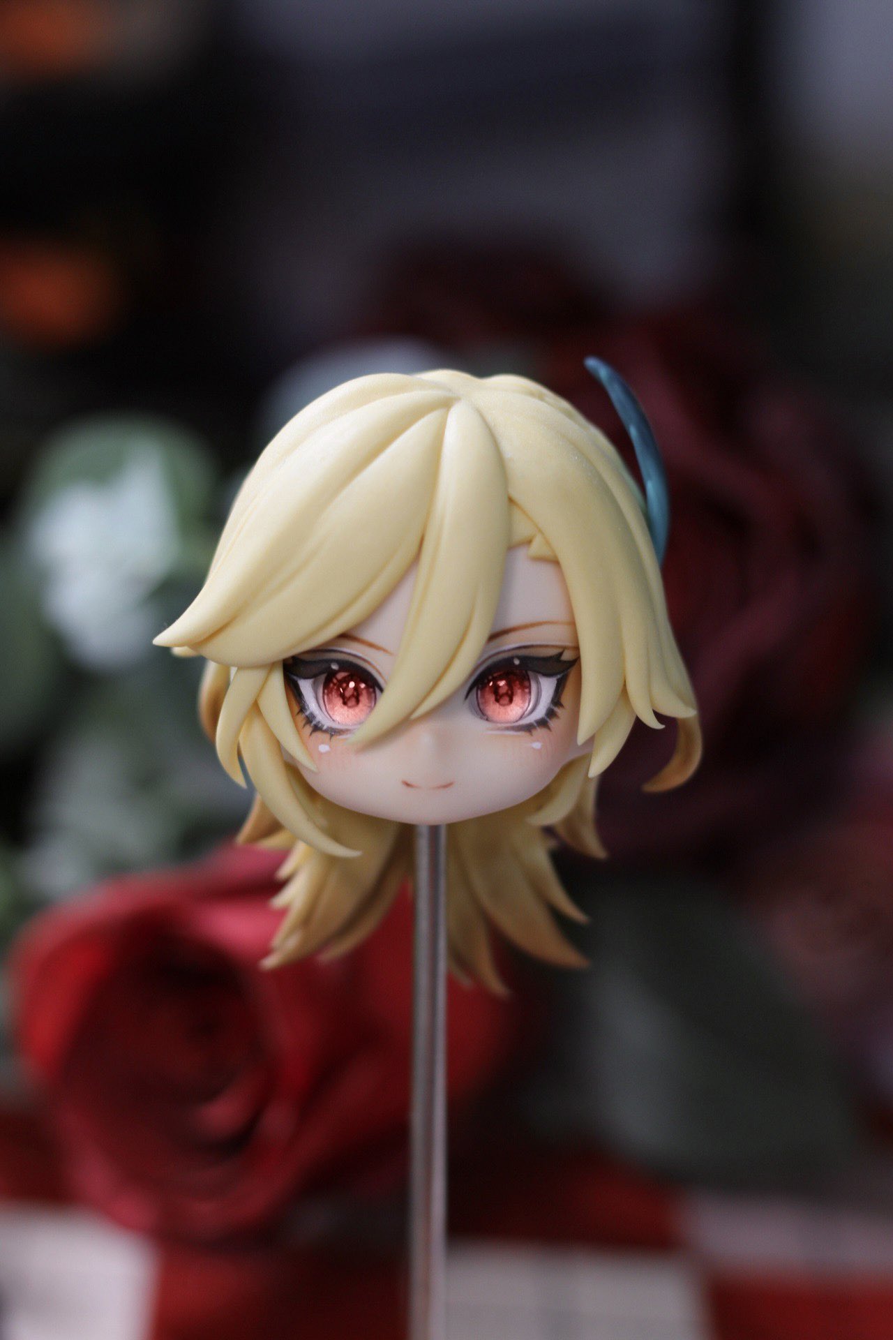 a closeup image of a handmade anime figurine. the figure is a blonde woman with red eyes and a smile. the figurine is made of plastic and has been hand painted with vibrant colors. she has a gentle expression on her face.