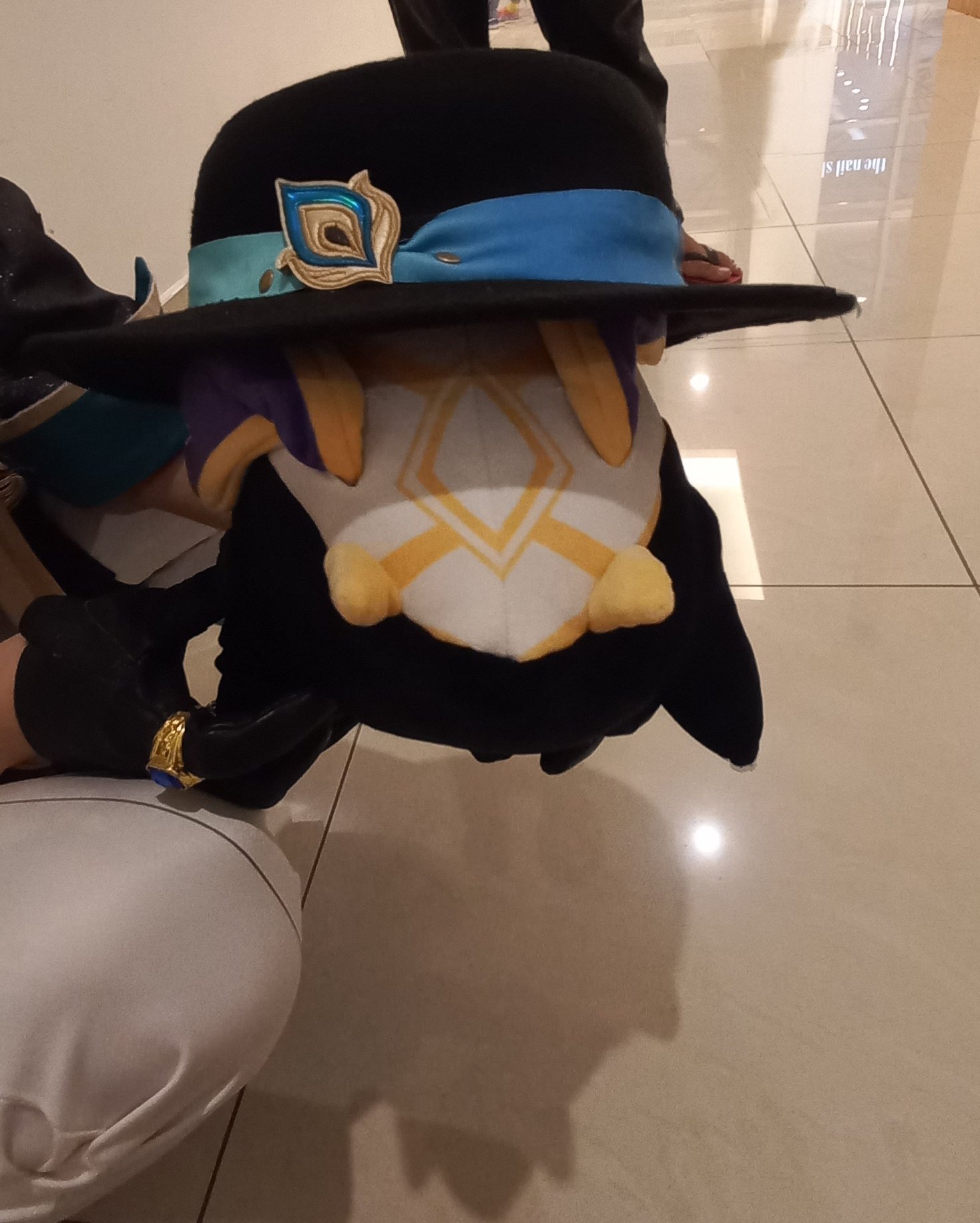 a black and blue hat with a gold feather sits on a white plush toy. the hat is covering most of the toy's head. the toy is sitting on a tan tile floor.