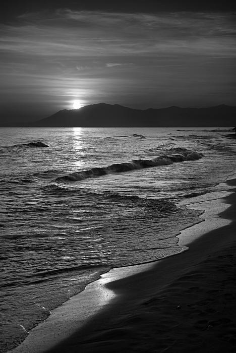 a black and white photo of a beach at sunset. the sun is setting over the horizon in the distance, casting a warm glow on the water and the sand. the waves are gently rolling onto the shore, and the mountains in the background are silhouetted against the sky. the sky is filled with clouds, and the overall mood of the photo is peaceful and serene.