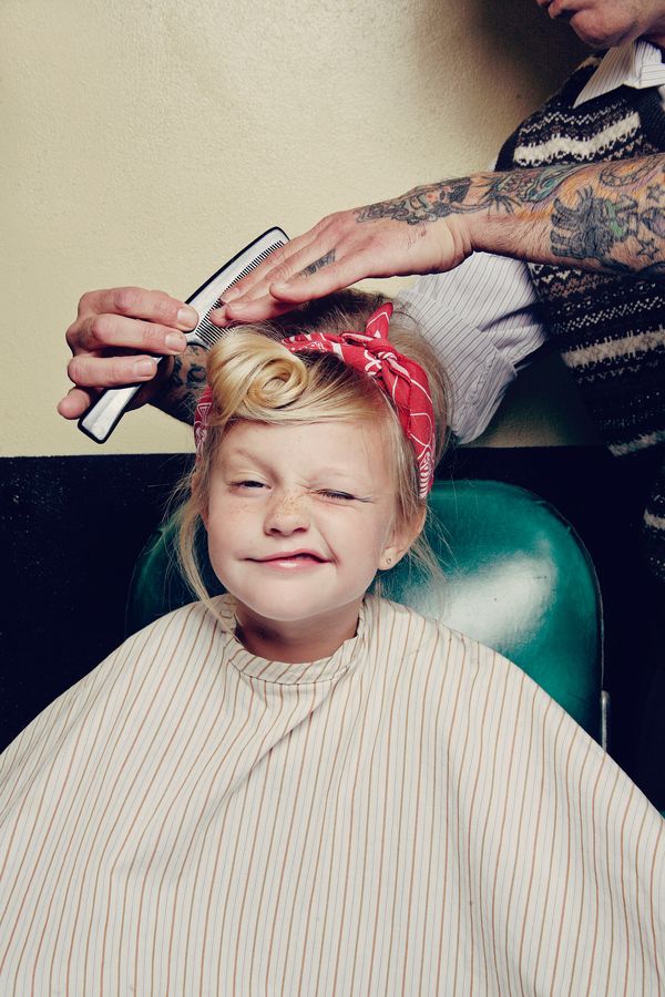 a young girl is getting her hair combed at a barber shop. she is wearing a red bandana in her blonde hair, and a striped barber cape. she is sitting in a green chair and winking at the camera. a barber with tattooed arms is combing her hair.
