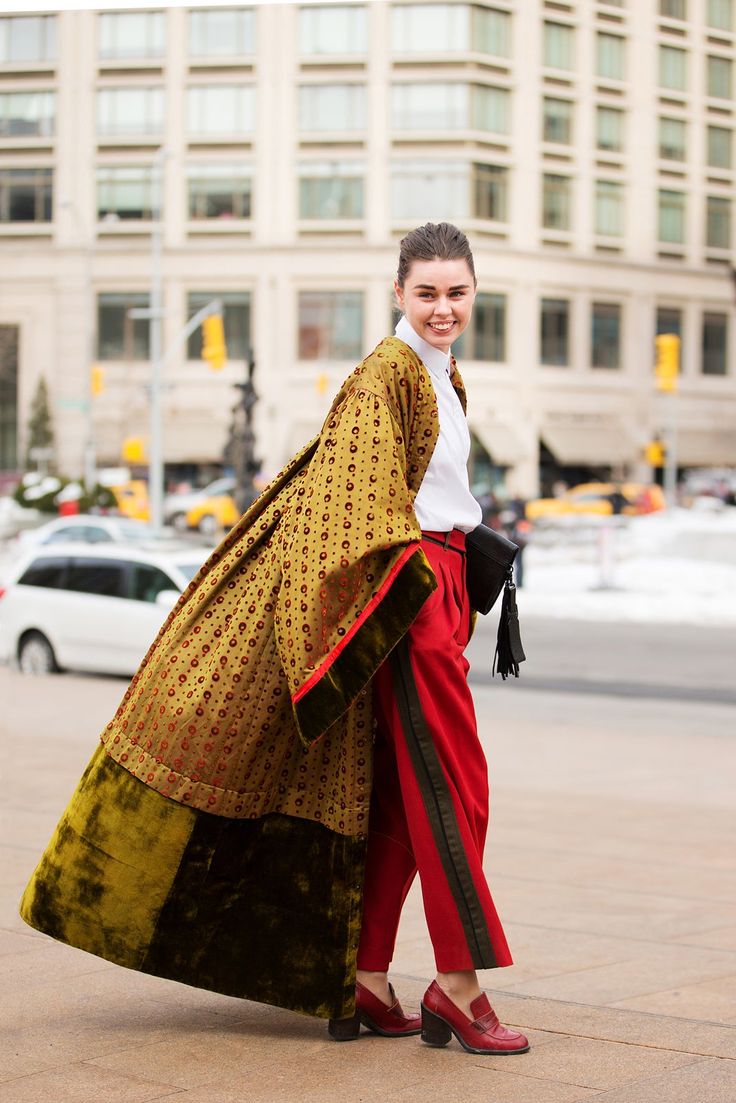 a stylish woman is posing on a city street, wearing a unique fashion ensemble. she's wearing red trousers with a dark green stripe down the side and matching red loafers with block heels. she has a white blouse on, and an olive green, patterned coat with a dark velvet trim that extends to the ankles, with a large olive green section toward the bottom, flows dramatically as she moves. she is also wearing a black bag with tassels. in the background, there are buildings, street traffic, and remnants of snow, suggesting an urban setting. the woman is smiling and looking at the camera.