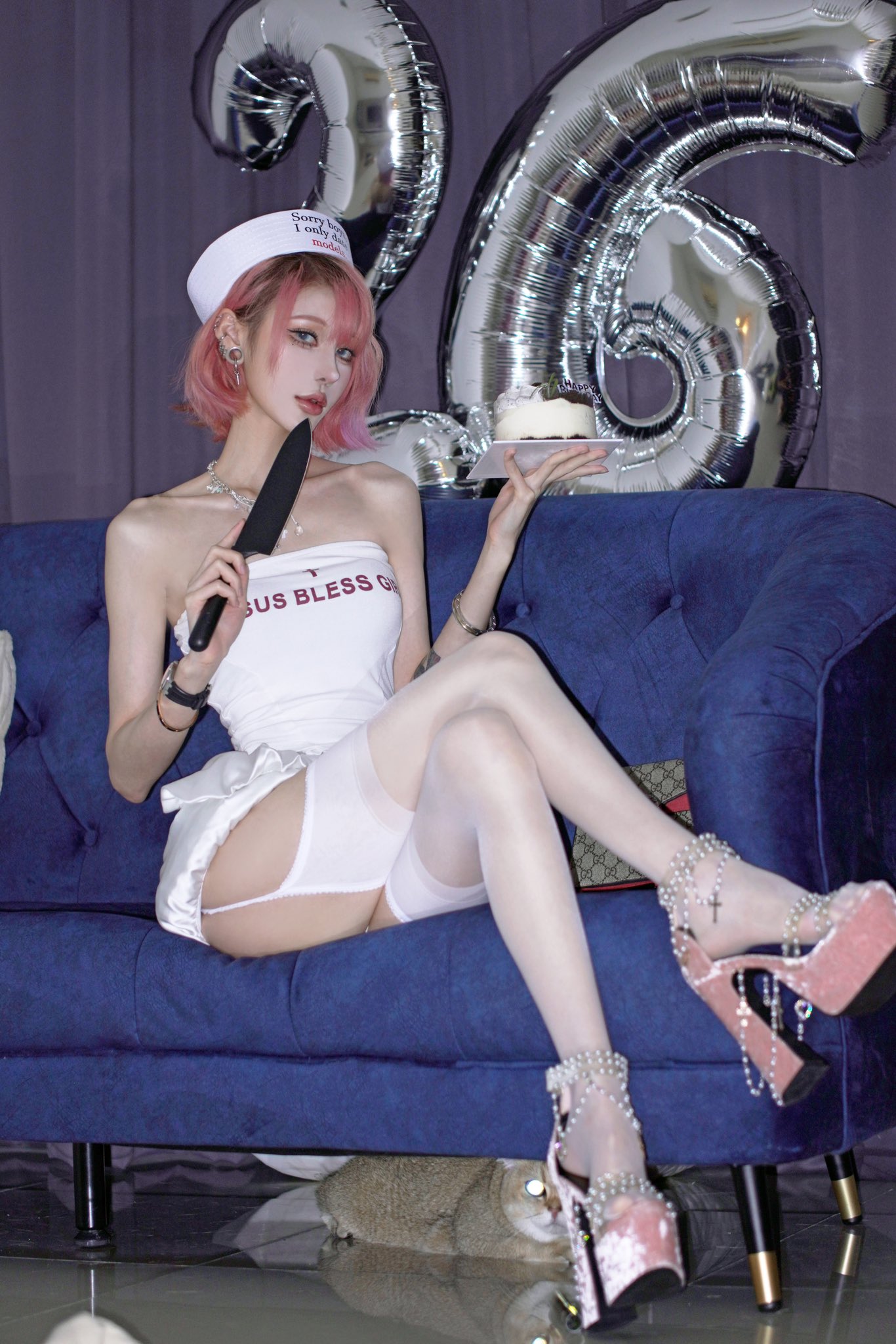 a young woman with short pink hair and a sailor hat is sitting on a blue couch. she's wearing a white dress, thighhigh stockings, and high heels. she's holding a knife and a piece of cake. there are silver number 26 balloons in the background.