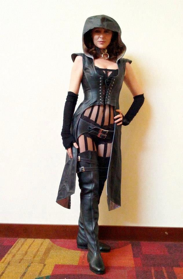 a woman in a black leather costume with a hood stands against a white wall with a red, orange, and brown patterned carpet floor. she is looking at the camera. she is wearing a black corset, boots, and a long, hooded, leather jacket. she is wearing a striped shirt and a belt around her waist.