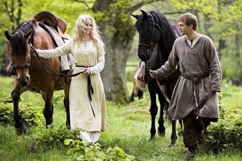 a man and a woman are walking through a forest with their horses. the woman is wearing a white dress and the man is wearing a brown tunic. the horses are brown and black. the forest is green and lush. the lighting is natural and bright. the image evokes a sense of love and romance.