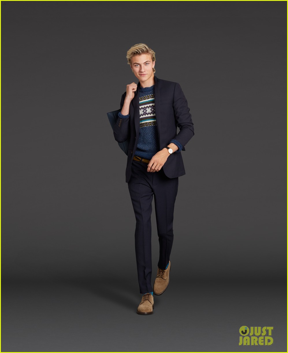 a blonde man is modeling a suit with a fair isle sweater and suede shoes. he is holding the blazer over his shoulder.