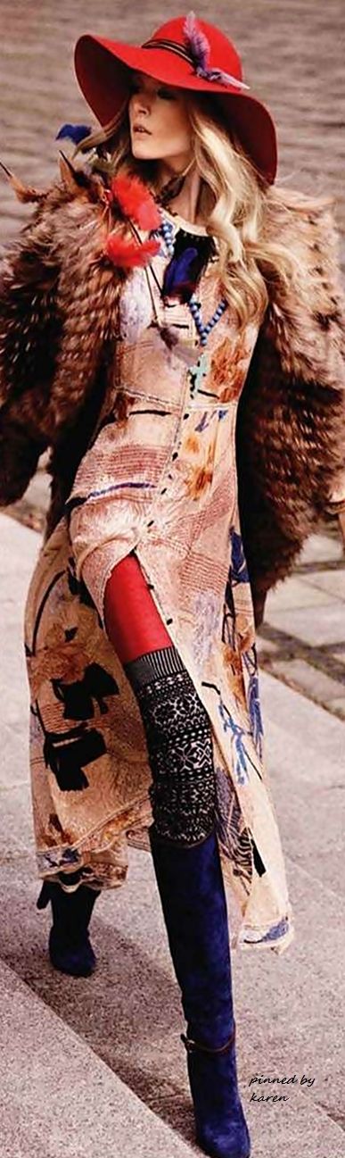 a young woman is wearing a stylish vintage outfit, including a red widebrimmed hat, a long brown fur coat, and a patterned dress with a long blue skirt. the model is posing with one hand on her hip, showing off her boots, which are blue suede and have a high heel. the background is a worn brick or concrete sidewalk.