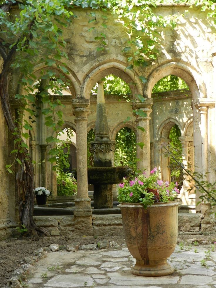 a sunlit courtyard with a stonepaved ground features an architectural structure with arches and columns covered in ivy, framing a fountain in the center. a large ornate pot filled with pink and green plants sits in the foreground, while other potted plants and foliage are scattered around the courtyard, adding to the serene and overgrown atmosphere. the stone walls and details of the architecture suggest an aged and classic style.