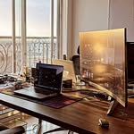 the image shows a modern and bright home office workspace.  a large, curved computer monitor takes center stage on a sleek, wooden desk. a laptop is open and positioned in front of the monitor, suggesting active work.  the desk is wellorganized with minimal clutter. natural light streams in from a large window overlooking a city view, contributing to the bright and airy atmosphere. the overall impression is one of a clean, efficient, and stylish workspace.