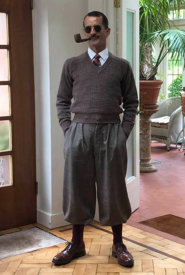 a man in vintage 1920s1930s style clothing stands in a hallway. he's wearing a brown vneck sweater, highwaisted pleated trousers, burgundy oxfords, and dark socks. he's also sporting a pipe, sunglasses, and a subtle mustache. the background shows a wooden floor, a doorway, and a conservatory with plants visible. the overall style is sophisticated and elegant.