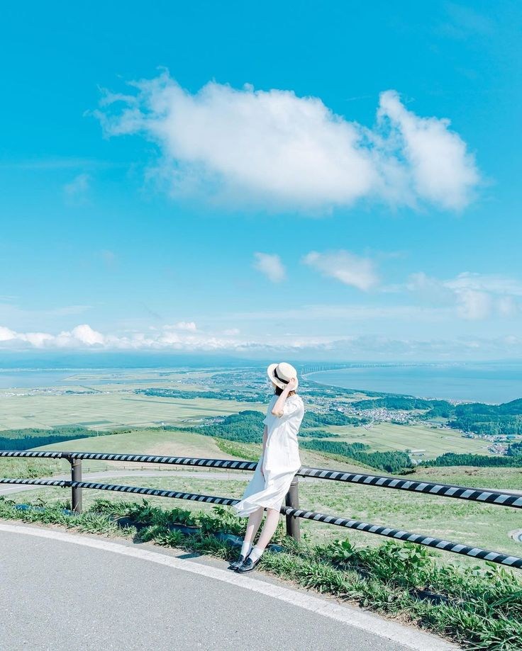 a woman in a white dress and hat stands leaning against a railing at a viewpoint, looking out at a vast landscape. the scene includes a blue sky with fluffy white clouds, green fields and distant buildings, with a road curving in the foreground. the image captures a sense of serenity and appreciation of natural beauty.
